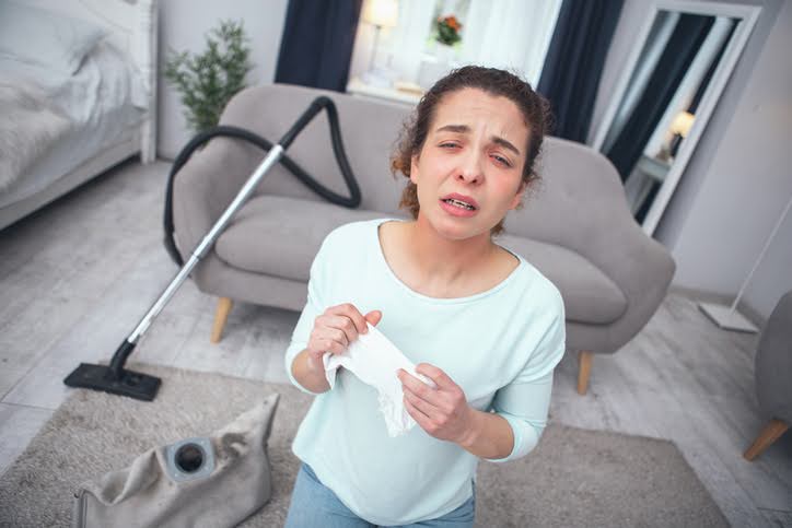 Woman about to sneeze holding a tissue in her hands with her vacuum laying behind her in the living room on her area rug.